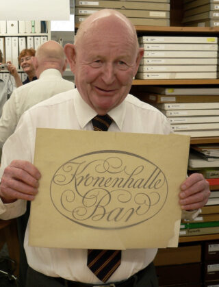 Ein älterer Mann in Hemd und Krawatte hält ein Schild mit der Aufschrift „Kronenhalle Bar“ in einem Raum voller Regale, Akten und Kisten.