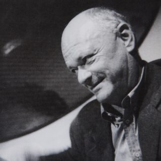 Schwarzweißfoto eines Mannes mittleren Alters mit Glatze, der ein Jackett trägt und mit einem leichten Lächeln zur Seite blickt.