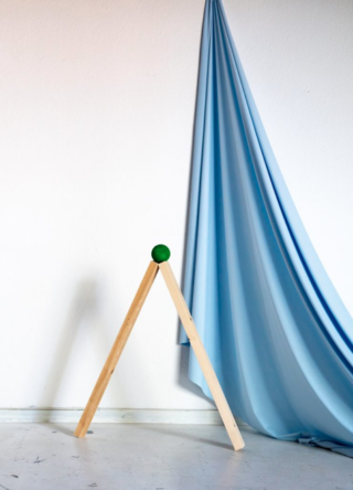 Zwei Holzbretter bilden ein umgedrehtes „V“, auf dem ein grüner Ball balanciert, neben einer mit fließendem hellblauem Stoff behangenen Wand.