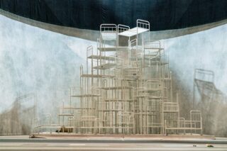 Eine große Skulptur aus gestapelten Metallbettrahmen ist in einer geräumigen Innenumgebung vor einem geschwungenen, schlichten Hintergrund stufenweise angeordnet.