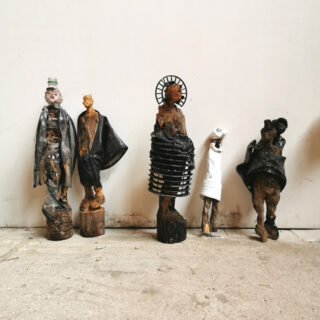 Fünf abstrakte, handgefertigte Figuren aus verschiedenen Materialien stehen in einer Reihe vor einer schlichten weißen Wand auf einem Betonboden.