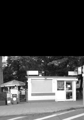 Schwarzweißfoto eines kleinen geschlossenen Kiosks mit einem Fenster mit Fensterläden, einem Regenschirm, einem Mülleimer und Bäumen im Hintergrund.