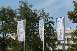 Drei vertikale Banner mit Texten in Französisch und Arabisch stehen vor einem modernen Gebäude mit Bäumen im Hintergrund unter einem klaren Himmel.