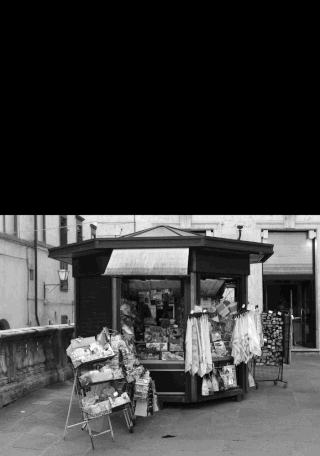 Auf einem gepflasterten Platz vor den Gebäuden steht ein kleiner Straßenkiosk mit Zeitungen, Zeitschriften und Postkarten.