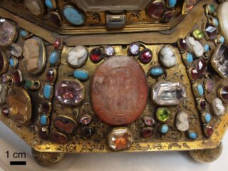 Eine Detailansicht eines kunstvollen, goldfarbenen Objekts, das mit Edelsteinen, Kameen und einem zentralen ovalen, geschnitzten Stein verziert ist. In der unteren linken Ecke befindet sich eine 1-cm-Skala als Referenz.