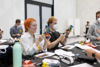 Menschen sitzen an einem Tisch und bauen kleine Roboter zusammen. Werkzeuge und Teile liegen verstreut auf der Tischplatte. Einige Teilnehmer tragen Masken und Sicherheitsetiketten.