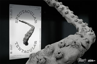 Eine graue, strukturierte Tentakelskulptur steht vor einem Poster mit der Aufschrift „Spectacular Tentacular“ in einem minimalistischen Schwarz-Weiß-Design.