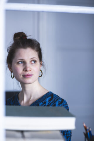 Eine Frau mit braunem Haar zu einem Dutt, einem blau gemusterten Pullover und Creolen trägt, schaut zur Seite, während sie drinnen neben einem Stapel Bücher sitzt.