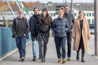 Sechs Erwachsene gehen gemeinsam im Freien auf einem gepflasterten Weg spazieren, gekleidet in legere und halbformelle Herbst- oder Winterkleidung, mit Gebäuden und Bäumen im Hintergrund.