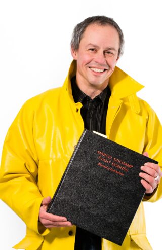 Ein Mann in einem gelben Regenmantel hält ein großes schwarzes Buch mit dem Titel „Marcel Duchamp Étant donnés“ und lächelt vor einem weißen Hintergrund in die Kamera.