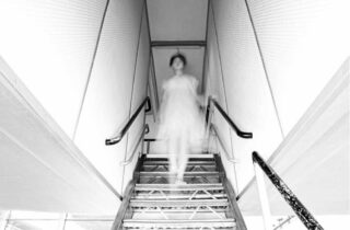 Eine verschwommene Gestalt in einem hellen Kleid steigt eine schmale Treppe mit Geländer hinab und erzeugt auf einem kontrastreichen Schwarzweißfoto einen geisterhaften Effekt.