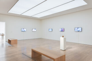 Eine minimalistische Kunstgalerie verfügt über sechs an der Wand montierte Bildschirme mit Bildern, einen zentralen Sockel mit einer Glasvitrine und zwei Holzbänke auf einem Hartholzboden.