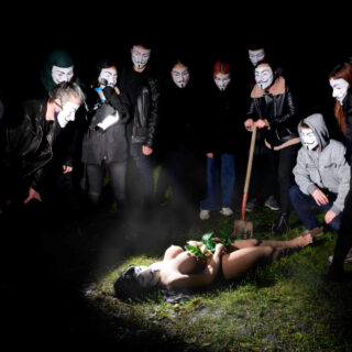 Eine Gruppe von Menschen mit Guy-Fawkes-Masken umringt eine nackte Person, die nachts im Gras liegt und Pflanzen hält; eine Person hält eine Schaufel.