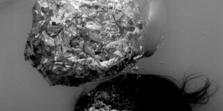 Der Kopf einer Person ist teilweise im Wasser eingetaucht. Auf der Wasseroberfläche sind Blasen und Wellen in Schwarzweiß zu sehen.