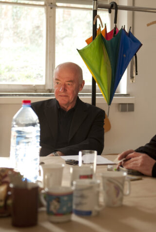 Ein älterer Mann im schwarzen Anzug sitzt mit Tassen, Gläsern und einer Wasserflasche an einem Tisch, hinter ihm hängt in der Nähe eines Fensters ein bunter Regenschirm.