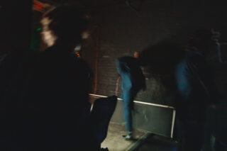 Drei Personen in einem schwach beleuchteten Raum; eine Person blickt auf eine dunkle Ziegelwand und ruht mit einer Hand darauf, während die anderen unscharf im Vordergrund zu sehen sind.