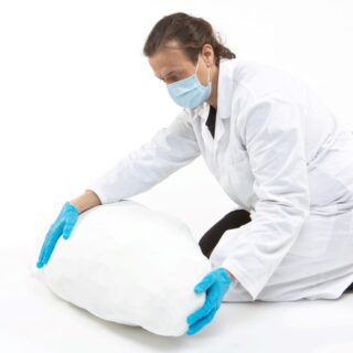 Eine Person mit Laborkittel, Maske und blauen Handschuhen kniet und handhabt vor einem weißen Hintergrund ein großes, weißes, unregelmäßig geformtes Objekt.