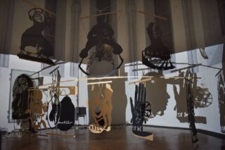 Mehrere abstrakte ausgeschnittene Formen und Silhouetten hängen von der Decke und werfen in einem schwach beleuchteten Raum komplexe Schatten auf den Boden und die Wände.