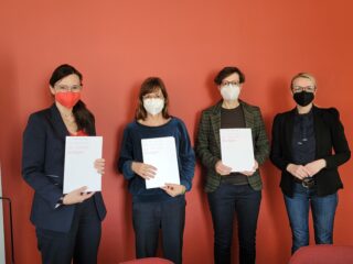 Vier Frauen stehen vor einer roten Wand, alle tragen Mundschutz und halten Dokumente mit der Aufschrift „Hochschule für Technik Stuttgart“ in den Händen.