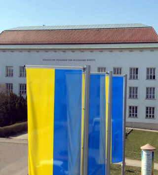 Vor einem großen weißen Gebäude mit rotem Dach stehen drei blau-gelbe Fahnen mit der Aufschrift „Staatliche Akademie der Bildenden Künste“.