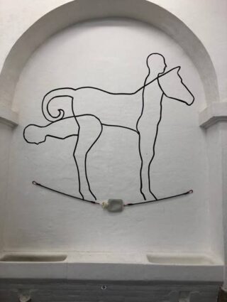 Eine minimalistische Drahtskulptur stellt die Umrisse einer menschlichen Figur und eines Pferdes dar, wobei sich ihre Formen auf einer weißen gewölbten Wand teilweise überlappen.