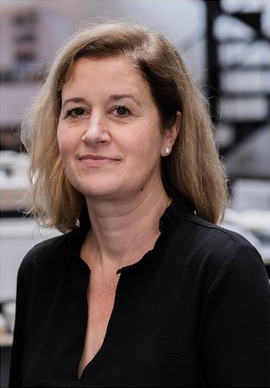 Eine Frau mit schulterlangem blondem Haar und einem schwarzen Oberteil steht mit neutralem Gesichtsausdruck im Innenbereich, im Hintergrund sind Büromöbel zu sehen.