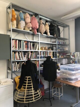 Auf Metallregalen finden sich Bücher, Hüte im historischen Stil auf Schaufensterpuppenköpfen, Kostümzubehör und Aufbewahrungsbehälter aus Kunststoff. Vor den Regalen sind auf zwei Schneiderpuppen historische Kleidungsstücke ausgestellt.