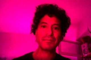 Eine Person mit lockigem Haar blickt in einem Raum mit hellrosa Licht in die Kamera. Im Hintergrund sind ein Fenster und einige Regale zu sehen.