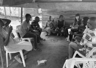 Eine Gruppe von Menschen sitzt im Kreis auf Plastikstühlen unter einem Unterstand und unterhält sich im Freien.