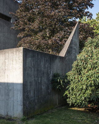 Betonwand mit eckigem Anbau neben dichten grünen Sträuchern und Bäumen, unter Tageslicht.