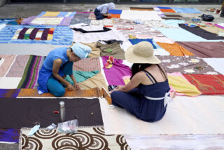 Zwei Personen sitzen auf großen, bunten Stoffbahnen, die auf dem Boden ausgebreitet sind, nähen und arbeiten mit Materialien; andere Stoffe und Materialien sind um sie herum verstreut.