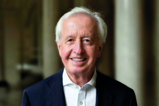 Ein älterer Mann mit weißem Haar, einem dunklen Jackett und einem weißen Hemd, lächelt in die Kamera, vor dem unscharfen Hintergrund von Säulen.