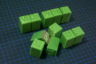 Grüne 3D-gedruckte Blöcke mit geprägten Wörtern sind auf einer blauen Gittermatte angeordnet; ein Block in der Mitte ist braun.