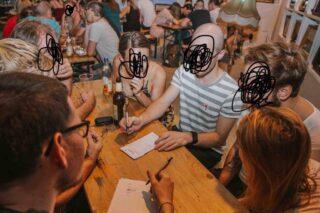 Sechs Personen sitzen mit Getränken und Notizblöcken um einen Holztisch und scheinen an einer Gruppenaktivität in einer Bar oder einem Café teilzunehmen. Ihre Gesichter sind durchgekritzelt.