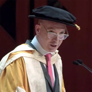 Eine Person in akademischer Kleidung und mit Mütze spricht an einem Podium und blickt zum Publikum.