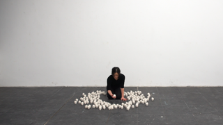 Eine schwarz gekleidete Person kniet auf einem gefliesten Boden und ordnet zahlreiche kleine weiße Objekte in einem Kreis vor einer schlichten weißen Wand an.