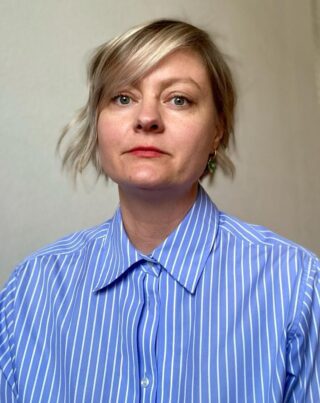 Eine Person mit kurzen, blonden Haaren und einem blau-weiß gestreiften Hemd mit Knöpfen blickt vor einem schlichten Hintergrund direkt in die Kamera.