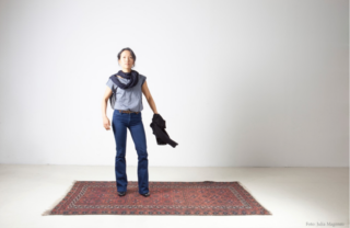 Eine Person steht auf einem gemusterten Teppich in einem minimalistischen Raum. Sie trägt Bluejeans und einen Schal und hält in der linken Hand eine schwarze Jacke.