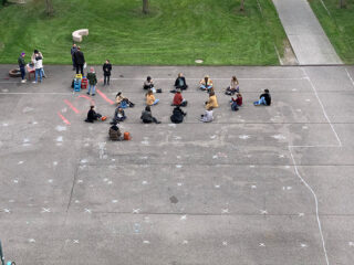 Eine Gruppe von Menschen sitzt mit Abstand zueinander in einem Kreis auf einer gepflasterten Fläche, die mit Kreide-X markiert ist, während andere in der Nähe auf dem Rasen stehen.