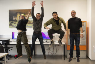 Vier Personen springen gleichzeitig in einer Büroumgebung vor Schreibtischen mit Computern herum; drei lächeln, einer blinzelt mitten im Gesicht.