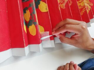 Eine Person bringt mit einer Pinzette einen kleinen weißen Fleck auf einem roten Papierobjekt mit aufgemaltem Fruchtmuster an.