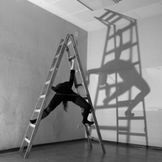 Eine Person in schwarzer Kleidung ist zwischen den Beinen einer offenen Leiter ausgestreckt und wirft in einem kahlen Raum den dramatischen Schatten einer verzerrten Gestalt auf die Wand hinter sich.