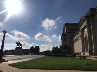 Unter einem sonnigen Himmel steht ein großes Steingebäude mit Säulen, mit einer Statue einer berittenen Figur und ein paar Autos, die entlang der Straße zu sehen sind.