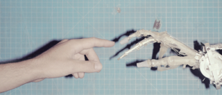 Eine menschliche Hand und eine transparente Roboterhand greifen vor einem blauen Gitterhintergrund aufeinander zu, wobei sich ihre Zeigefinger fast berühren.