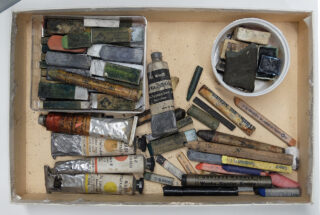 Eine Kiste mit verschiedenen gebrauchten Künstlerutensilien, darunter Ölfarbentuben, Pastellstifte, Bleistifte und Blöcke, die willkürlich auf einer beigen Oberfläche angeordnet sind.