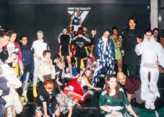 Eine bunt gemischte Gruppe von Menschen posiert in eklektischen, futuristischen Outfits in einem hell erleuchteten Raum. An der Wand hinter ihnen hängt ein Schild mit der Aufschrift „OWN THE REALITY“.