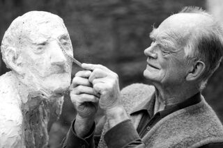 Ein älterer Mann formt eine Büste aus Ton und verwendet dabei ein Werkzeug, um das Gesicht der Figur mit konzentriertem Ausdruck zu formen.