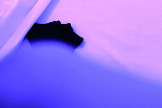 Ein schwarzes T-Shirt, teilweise mit einem weißen Laken bedeckt, über die Szene fällt ein blau-violettes Licht.