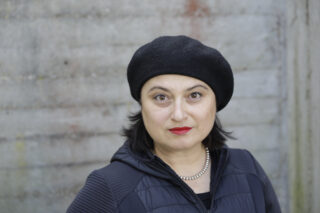 Eine Frau mit dunklem Haar, einer schwarzen Baskenmütze, einer schwarzen Jacke und einer Perlenkette steht vor einer Betonwand und blickt direkt in die Kamera.