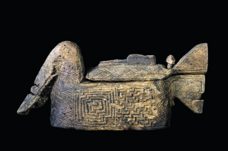 Eine geschnitzte Holzkiste mit geometrischen Mustern und einer darauf liegenden menschlichen Figur mit angewinkelten Knien und dem Kopf auf den Händen ruhend, vor einem schwarzen Hintergrund.